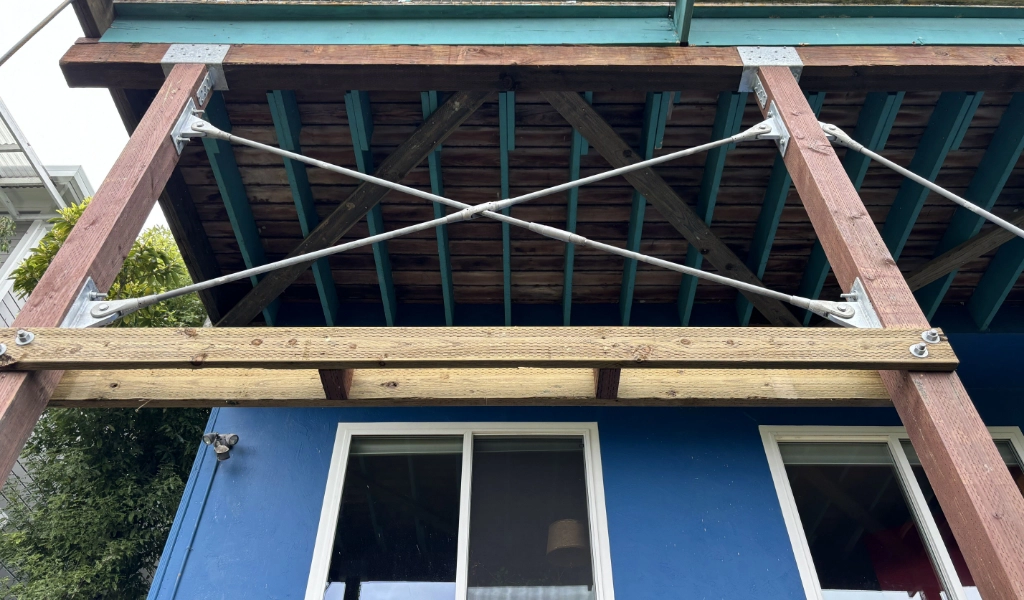 From below, a blue house showcases a wooden balcony, supported by metal cross-bracing and sturdy beams. Its exterior reveals two prominent windows, adding charm to this hillside retreat.