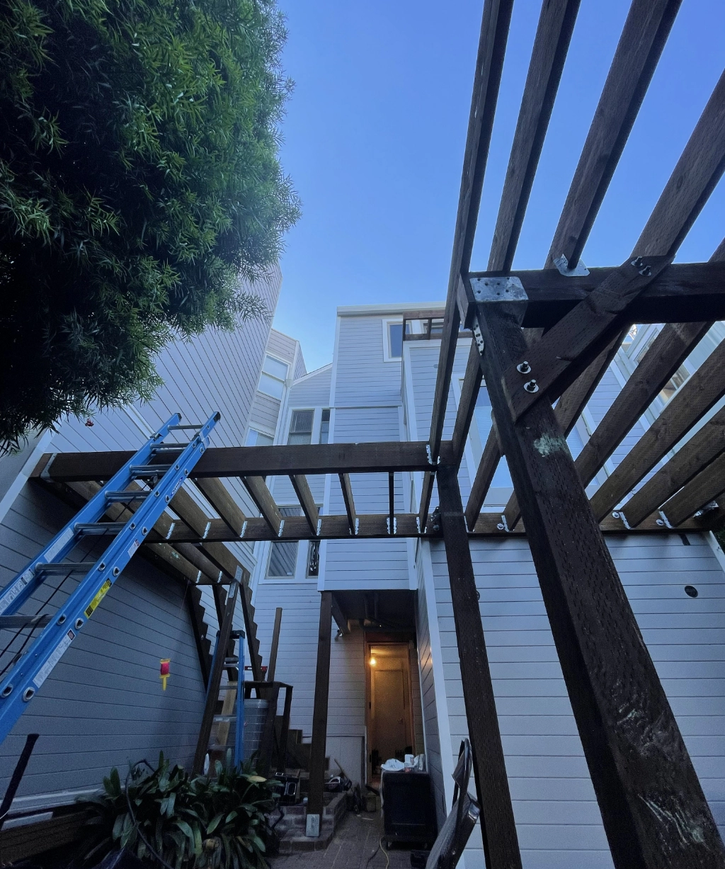 A wooden pergola is under construction with a blue ladder leaning against it. The building exterior, featuring white siding, stands proudly in the background, while a tree graces the scene on the left side.