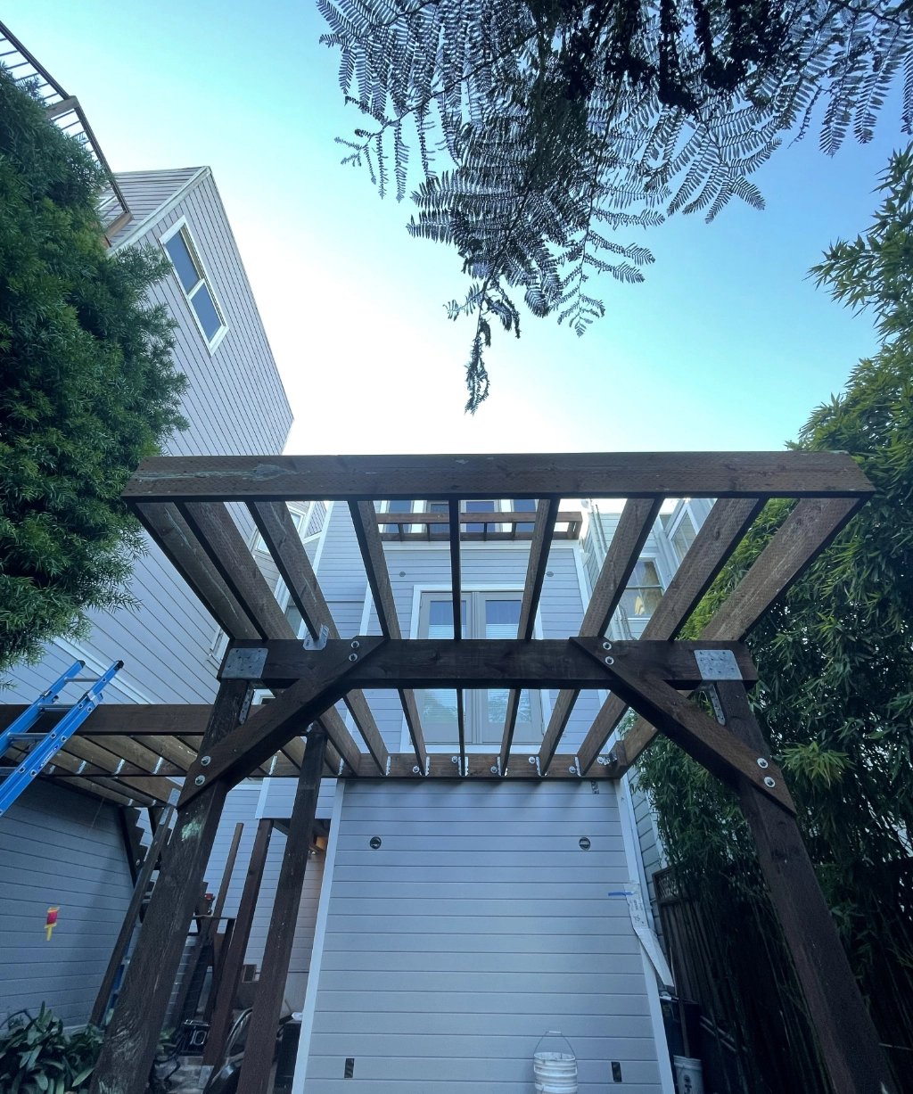 The wooden pergola structure graces the backyard of a modern home, framed by lush trees and vibrant vegetation. A blue ladder casually leans against the balcony, completing this picturesque scene.