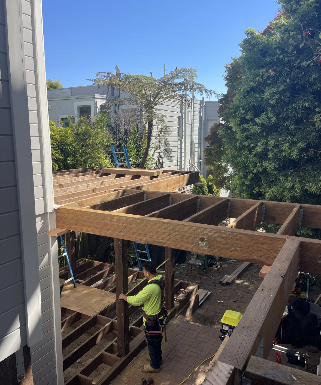 A construction worker skillfully builds a wooden deck structure between two buildings, surrounded by trees and a clear blue sky.