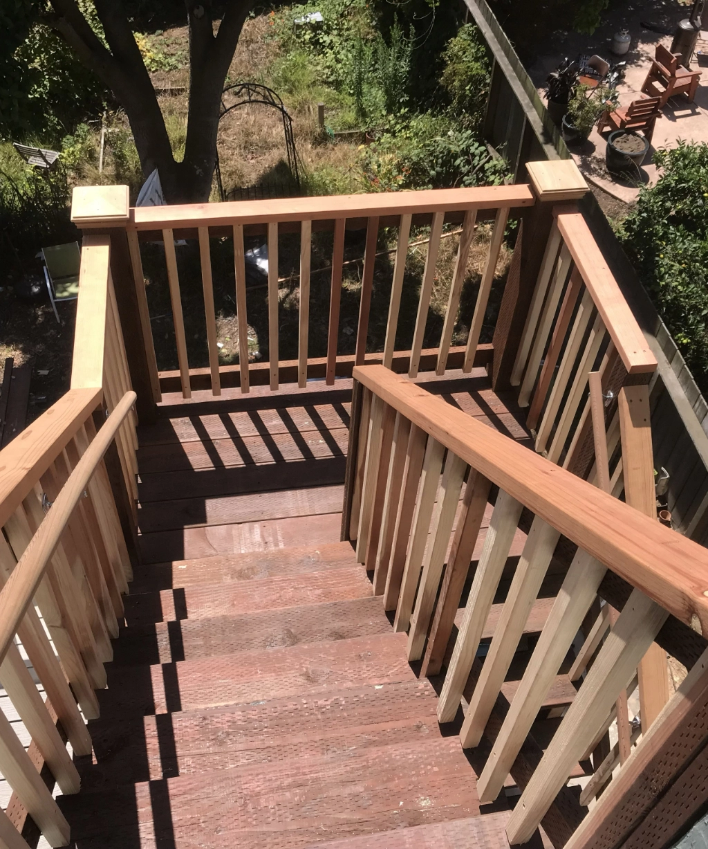 Wooden stairs with a railing gracefully lead down to a backyard. The area is surrounded by a wooden fence and lush trees, with some gardening tools and plants adding character to the serene setting.