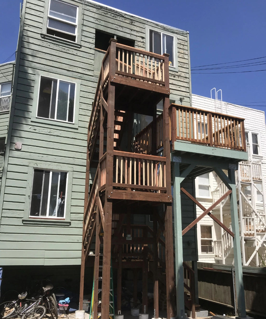 A wooden exterior staircase graces a green, three-story building, where bicycles rest nearby under a vibrant blue sky.