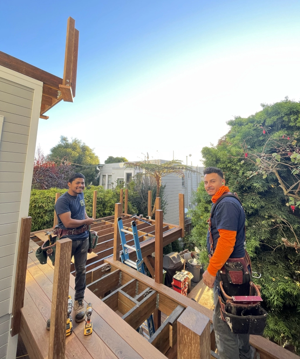 Two construction workers stand on an unfinished deck while smiling at the camera. The area is surrounded by lush greenery.