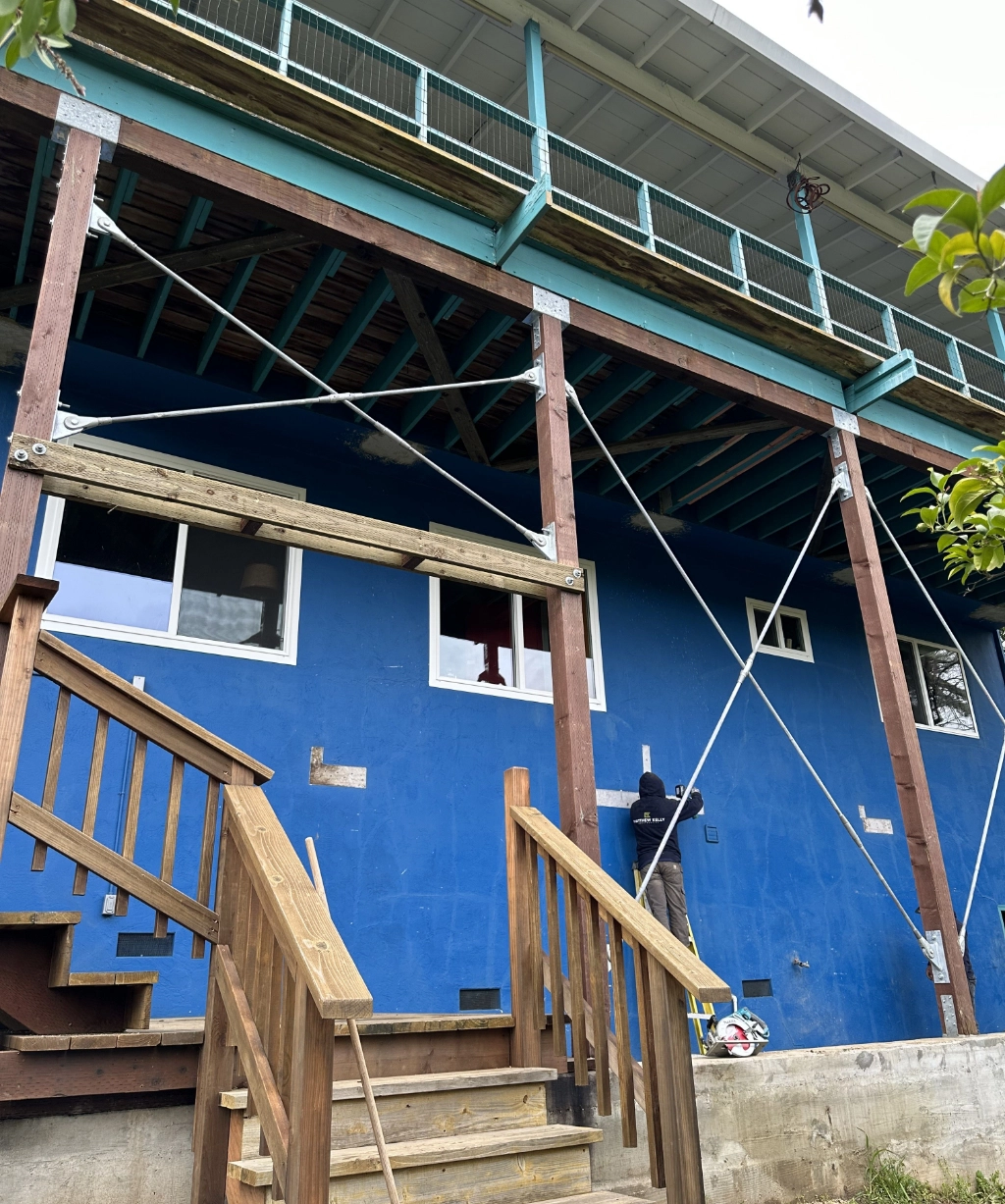 A person works on a blue exterior wall on a raised structure with wooden beams and cross-braces. A wooden staircase leads to a door on the left, offering an inviting entrance to this elevated retreat.