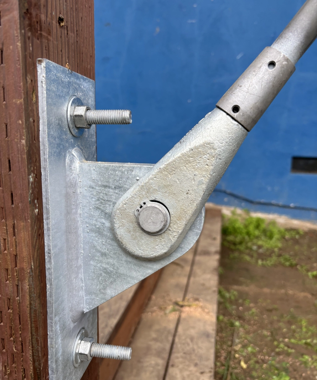 The metal hinge mechanism, securely bolted to a wooden post, stands adjacent to a pipe or rod.
