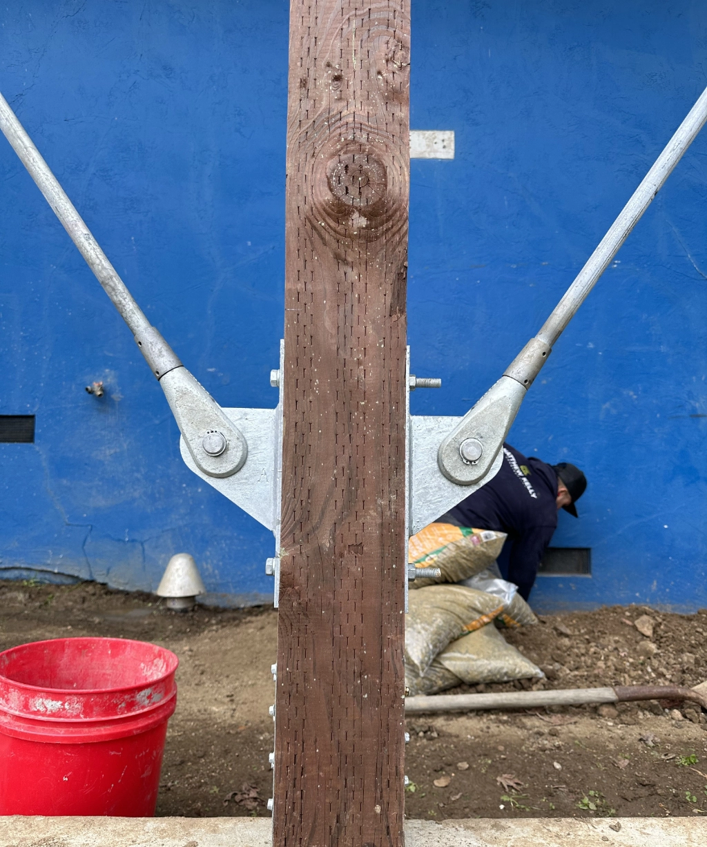 A wooden post with metal bracing stands in front of a blue wall; nearby, a person arranges bags beside a red bucket on the soil.