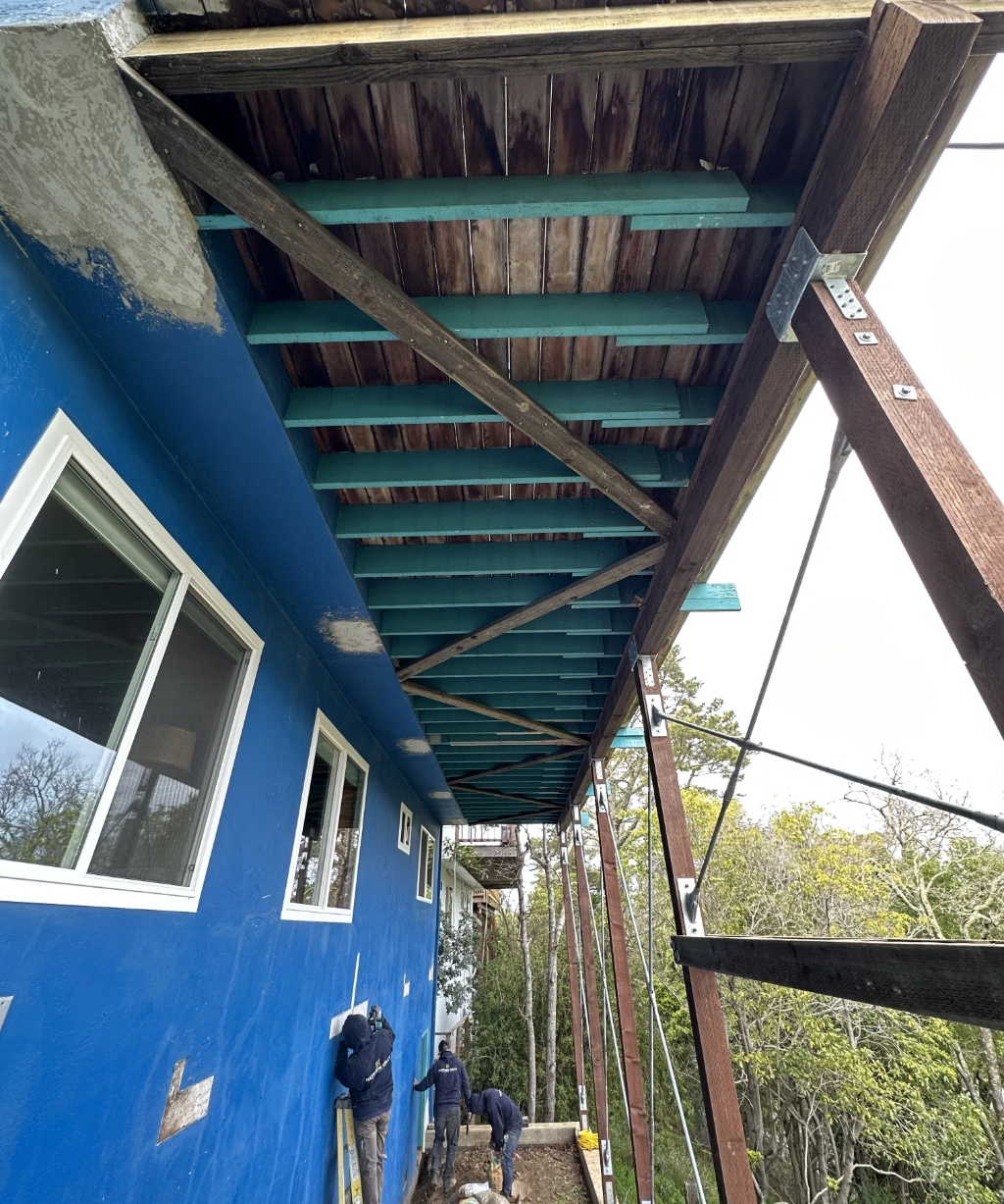 From beneath the deck of a blue building, you can see wooden beams and temporary support structures. Below, three people are hard at work.