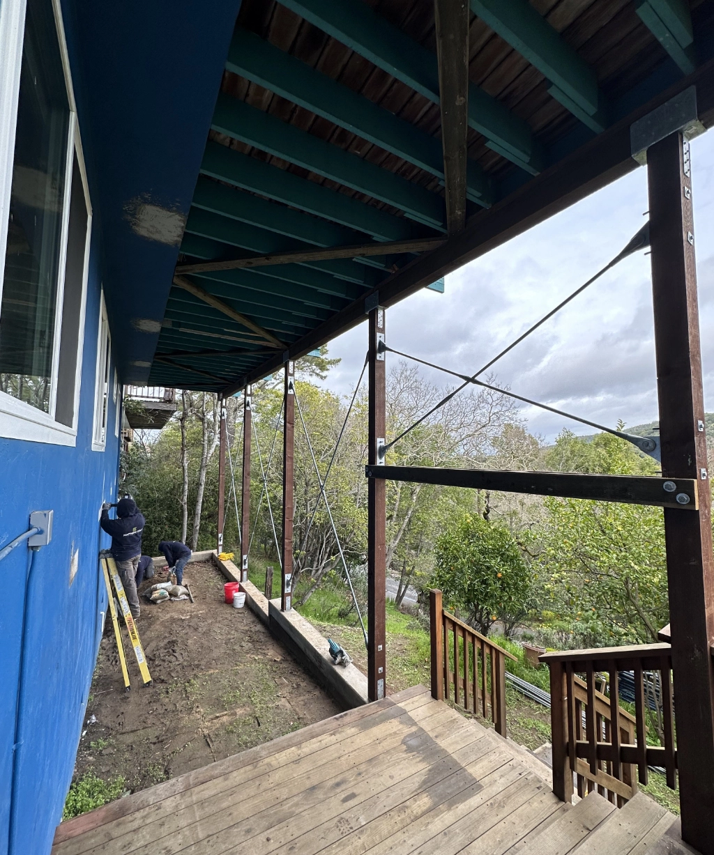 Construction workers reinforce the foundation of a blue house, using scaffolding. The scene includes trees, a gray sky, and a wooden staircase.