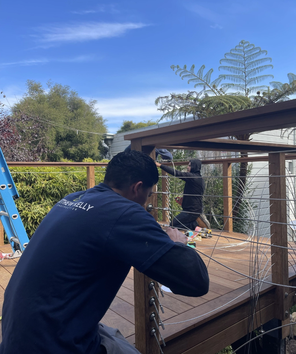 Two people are busy at work on a wooden deck. One is crouching to install wire railings, while the other stands near a ladder, surrounded by lush greenery.