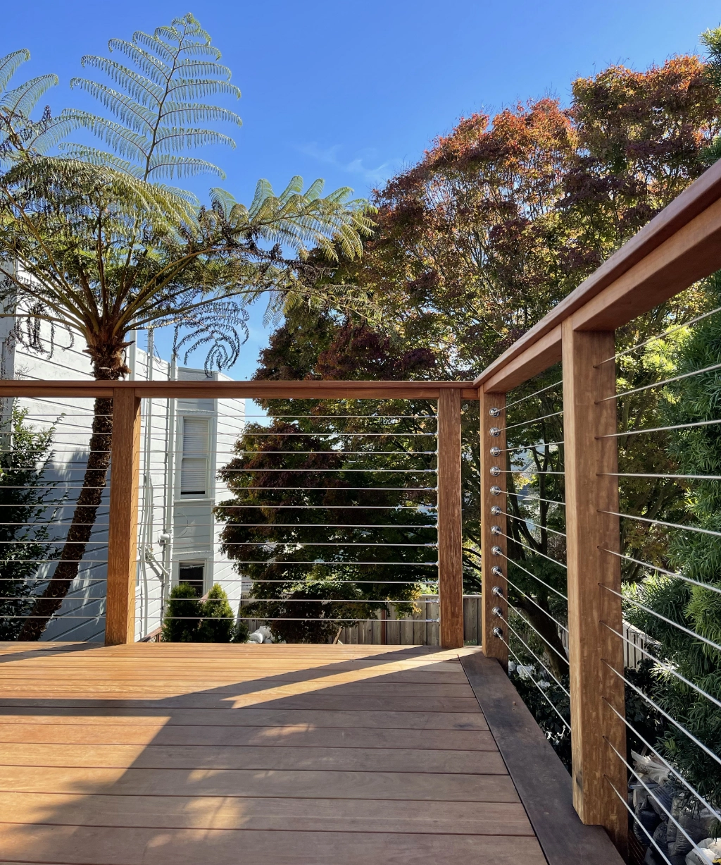 This wooden balcony with cable railing offers stunning views of trees and neighboring houses beneath a clear blue sky.