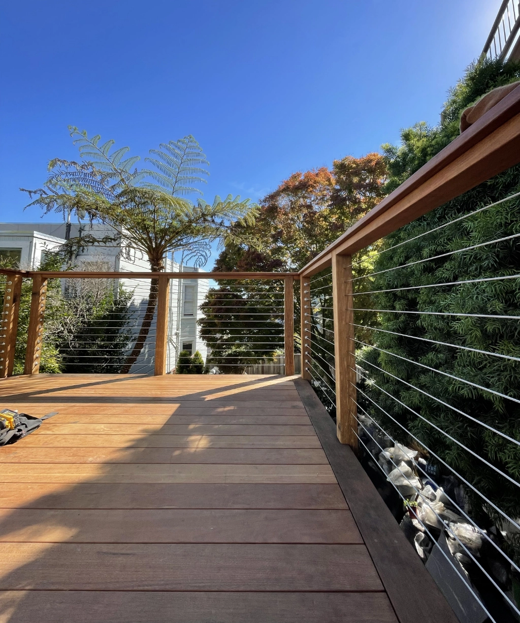 This wooden deck features sleek cable railing and is enveloped by lush trees and greenery under a clear blue sky.