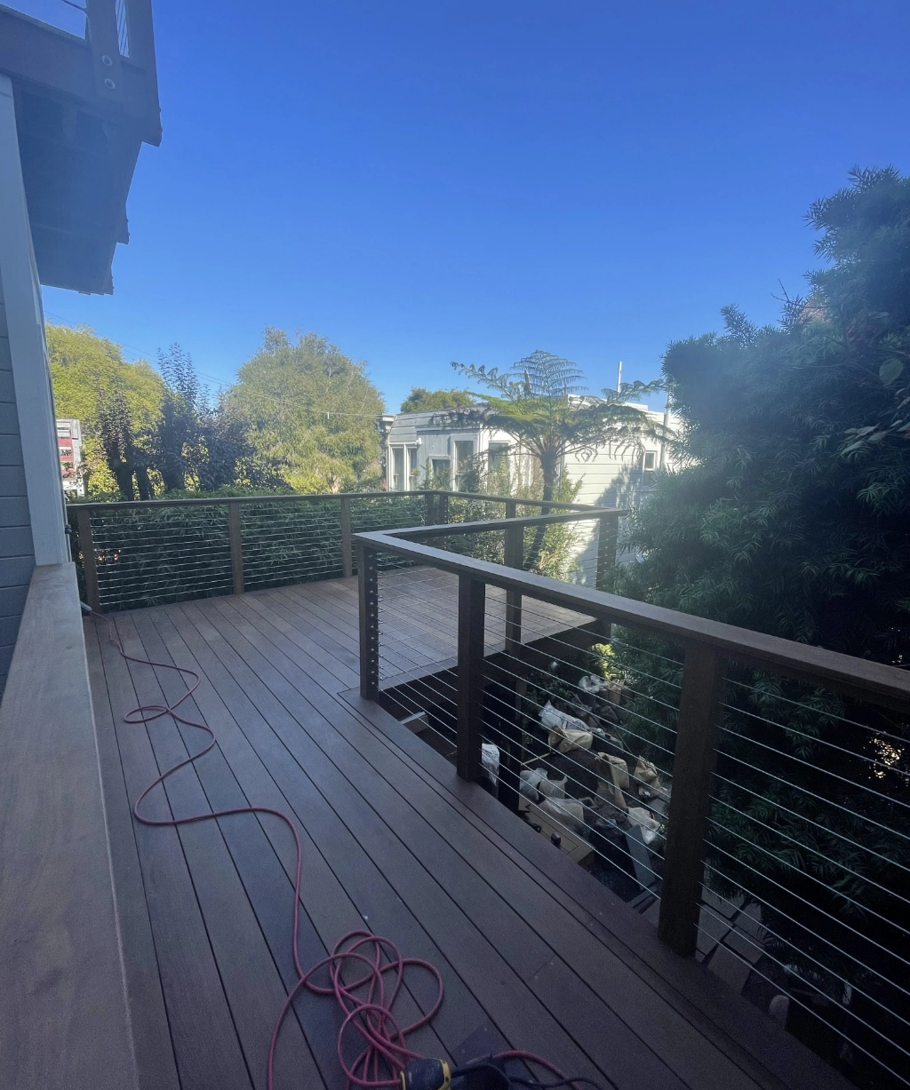 The wooden deck with railing and cable wires offers a stunning view of trees and the urban landscape; a red extension cord lies on the deck surface.