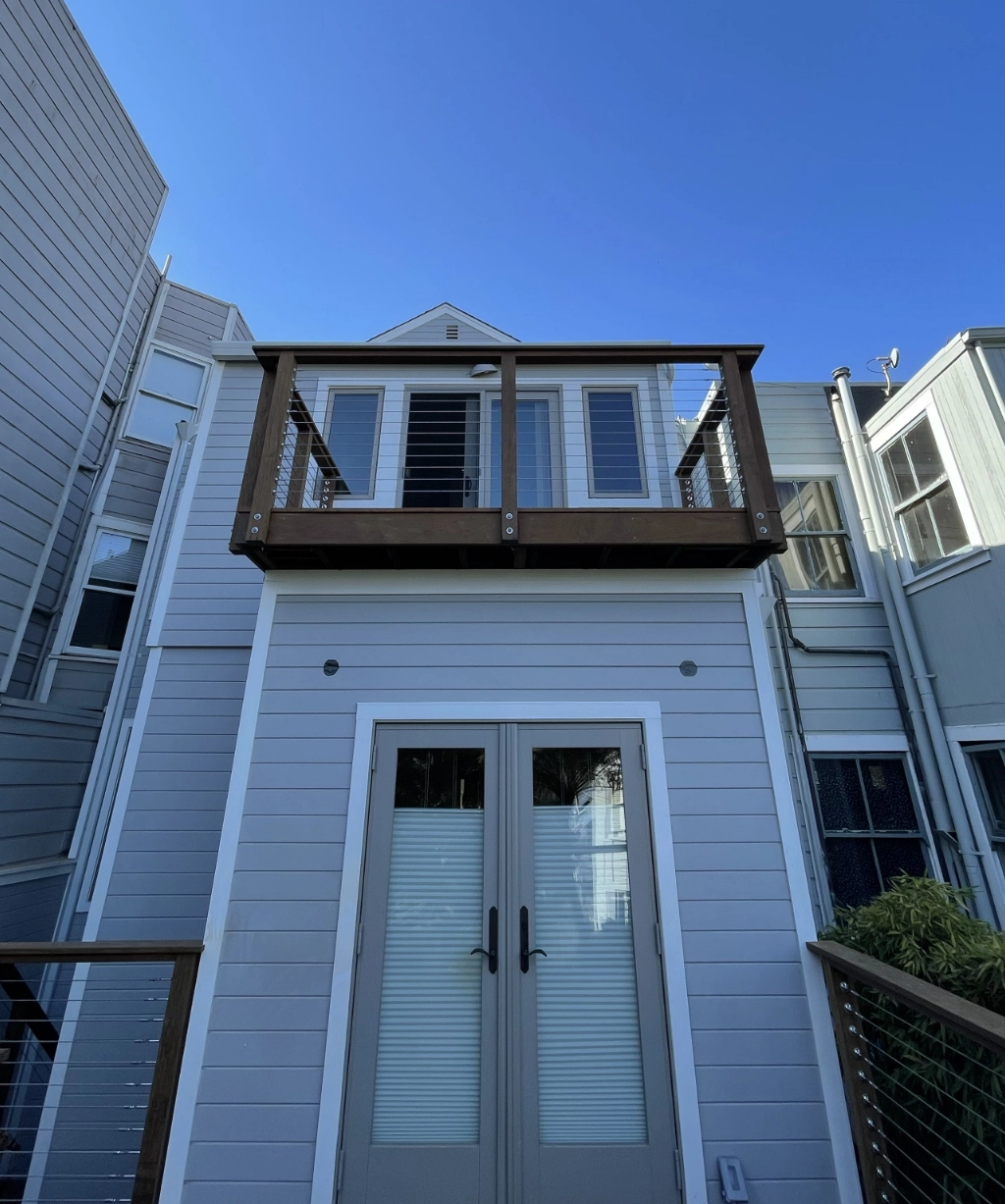 The two-story building boasts light gray siding and a charming wooden balcony with glass doors, all set against a clear blue sky.