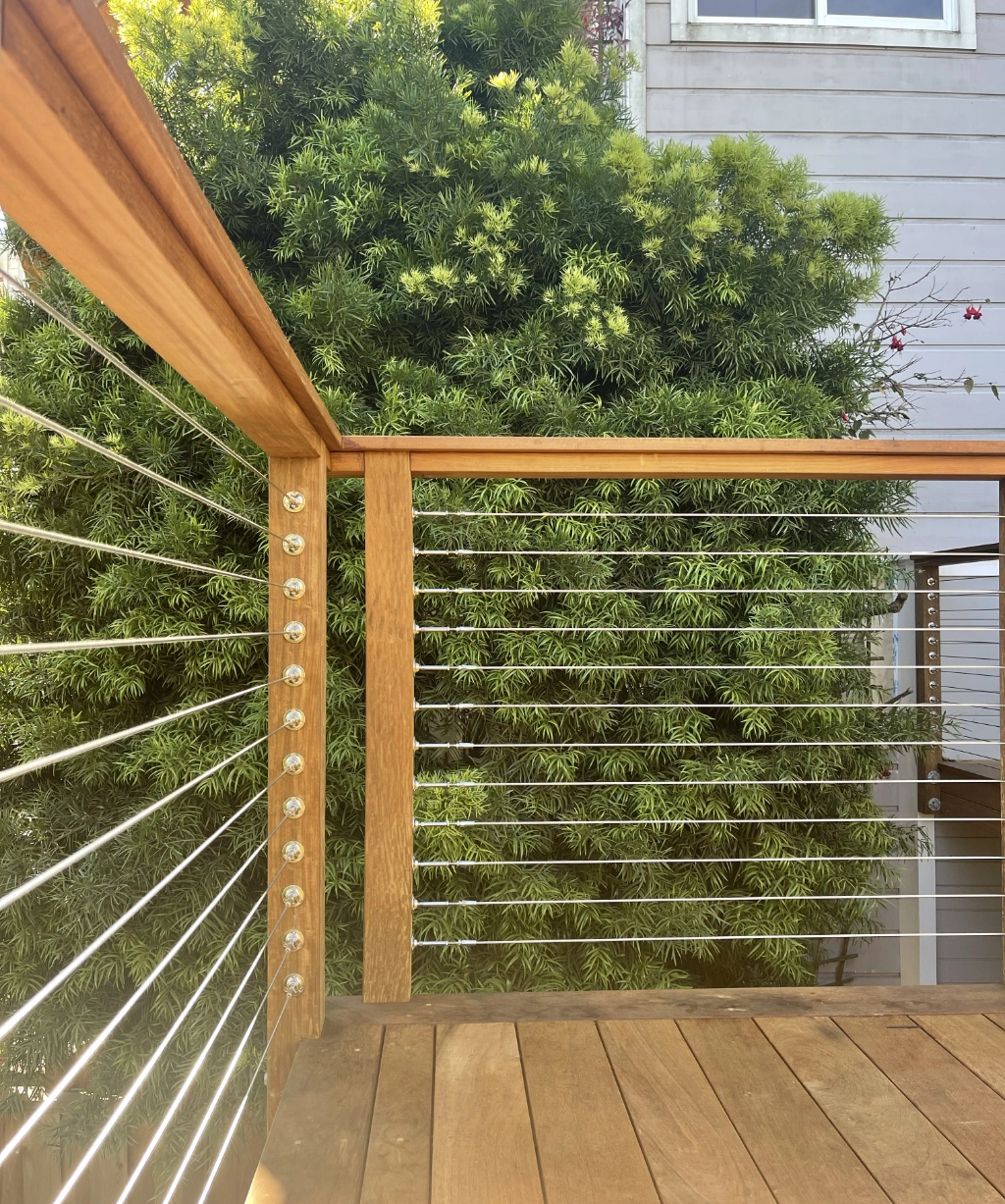 Nestled in picturesque surroundings, a wooden deck with a sleek metal railing offers a perfect vantage point to enjoy the adjacent lush green tree. In the background, a building wall subtly peeks through, adding architectural charm to this serene setting.