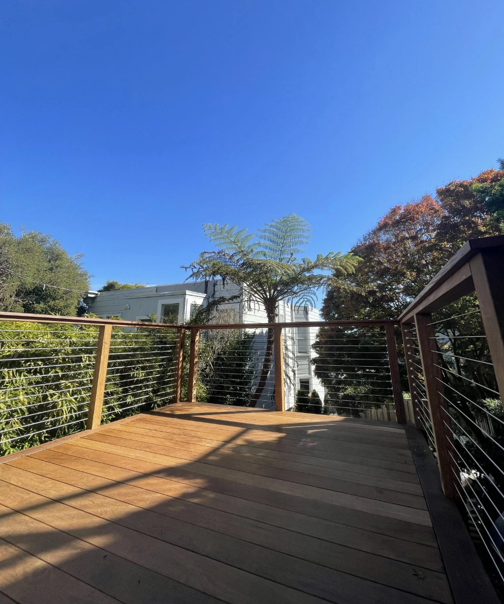 A wooden deck with a sleek metal railing provides a stunning view, overlooking lush trees and a pristine white building beneath the clear blue sky.