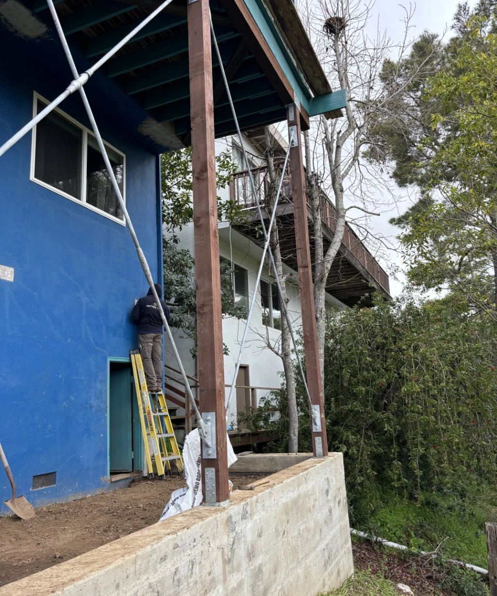 A person is standing on a ladder, tending to the blue exterior wall of a house. The structure is supported by metal braces and wooden beams, with a tree and another house nestled in the backdrop.