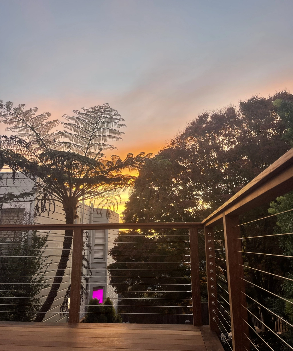 Sunset view from a wooden deck, with a railing in the foreground and trees silhouetted against the colorful sky in the background.