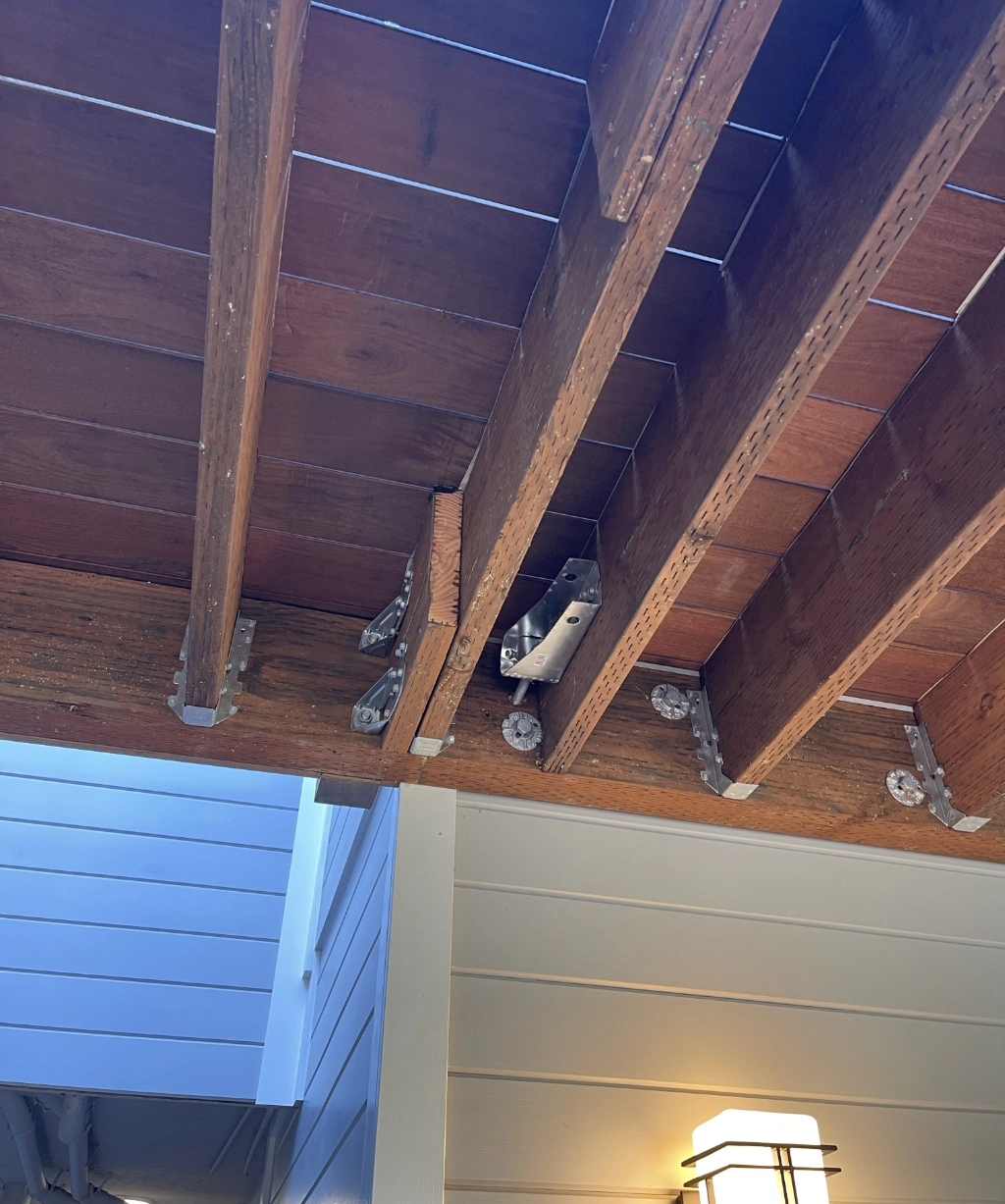Rustic charm comes alive with wooden ceiling beams supported by metal brackets, complemented by a light fixture on the wall below and adjacent white vertical siding.