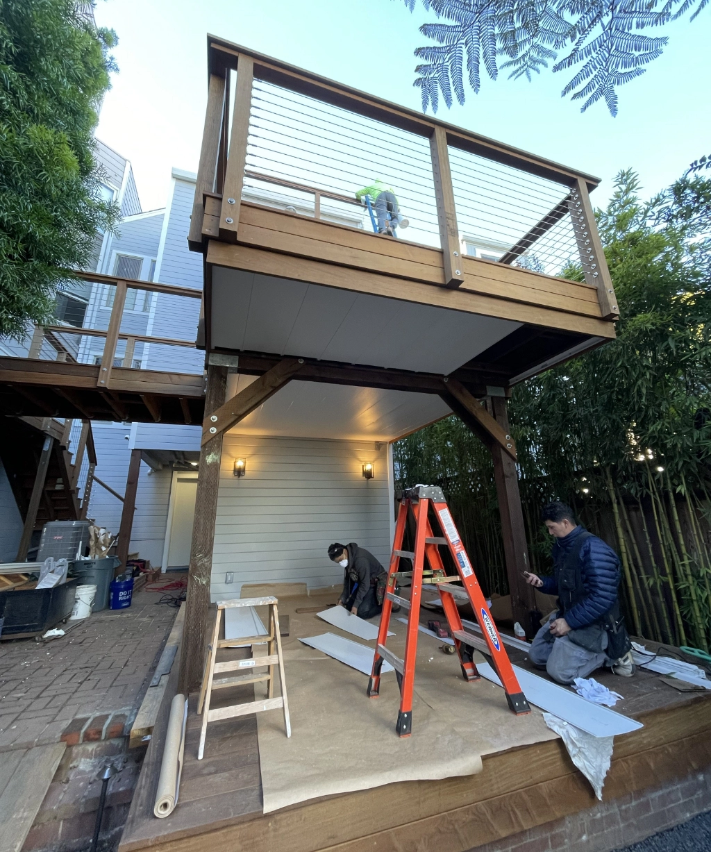 Two workers are at a job, installing under a wooden deck. One adjusts cables on the deck while the other uses tools on the ground. A red ladder and various materials surround them, with trees and a house providing a scenic backdrop.