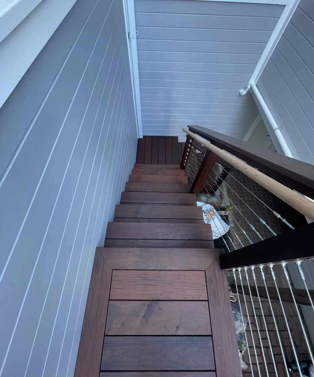 A wooden staircase with cable railing gracefully descends alongside a gray paneled wall. Some items are visible at the bottom of the stairs.
