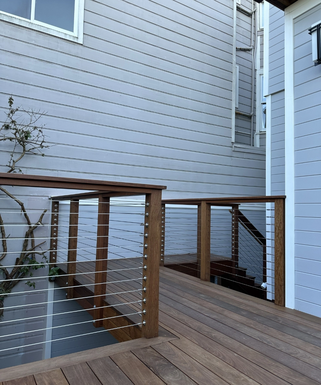 The wooden deck with cable railings, nestled against the house's light gray siding, features a staircase descending to a lower level. A lush plant adds charm on the left side.