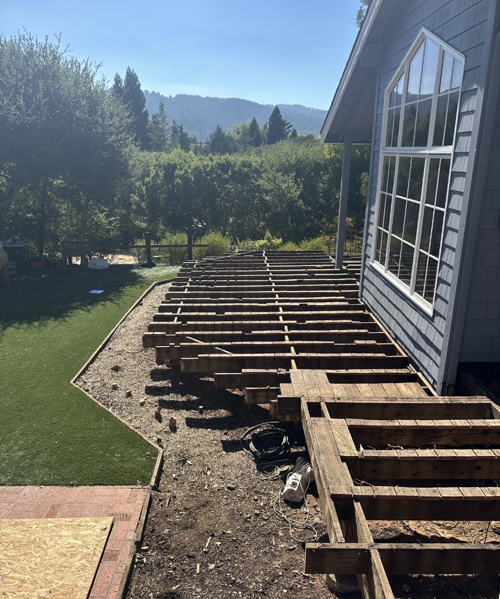 The wooden framework of a deck in progress is attached to a house, set against artificial grass. Majestic trees and rolling hills are visible in the background under a clear sky.
