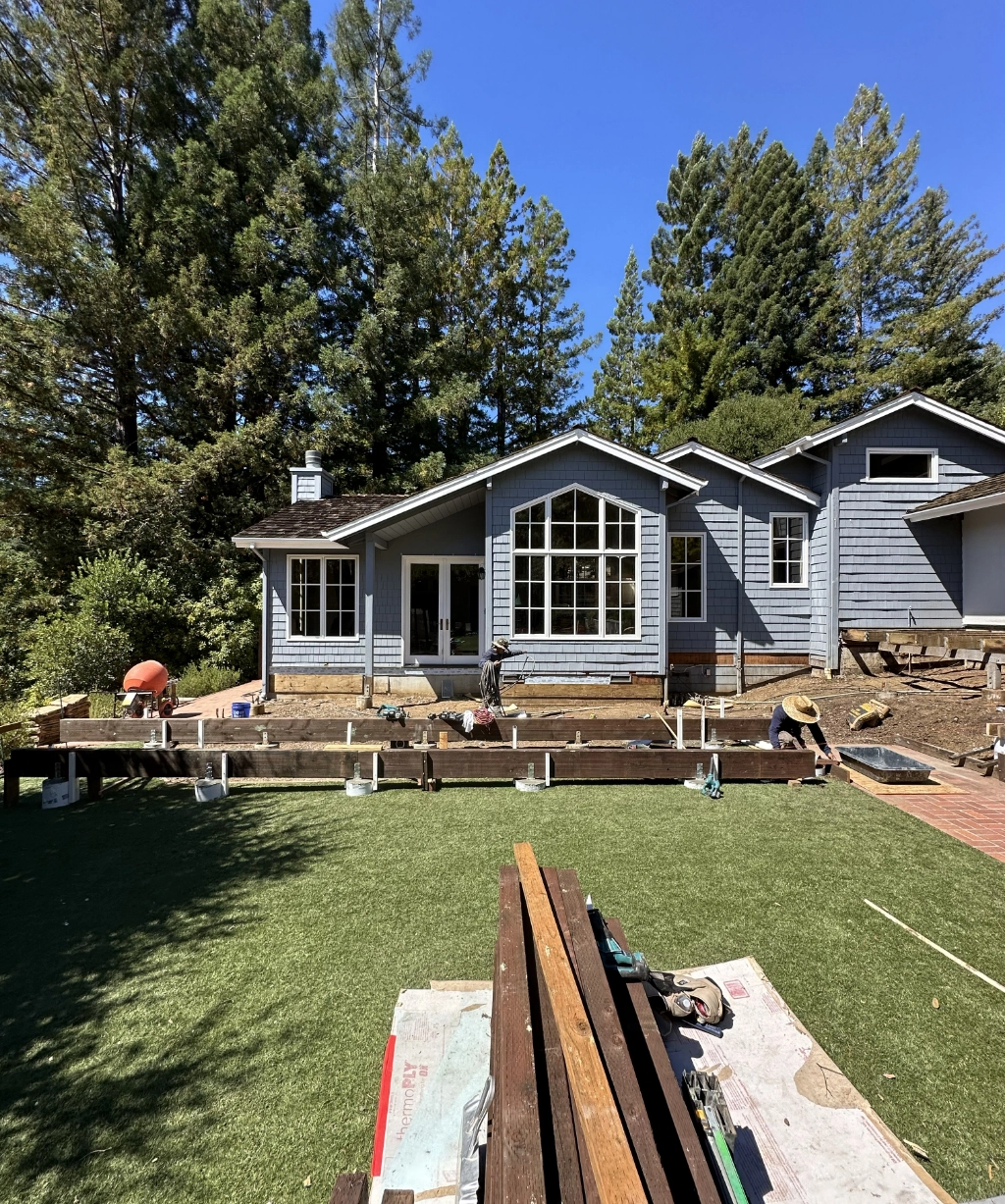 A house is under renovation, adorned with scaffolding and building materials scattered in the yard, all set against a backdrop of trees under a clear blue sky.