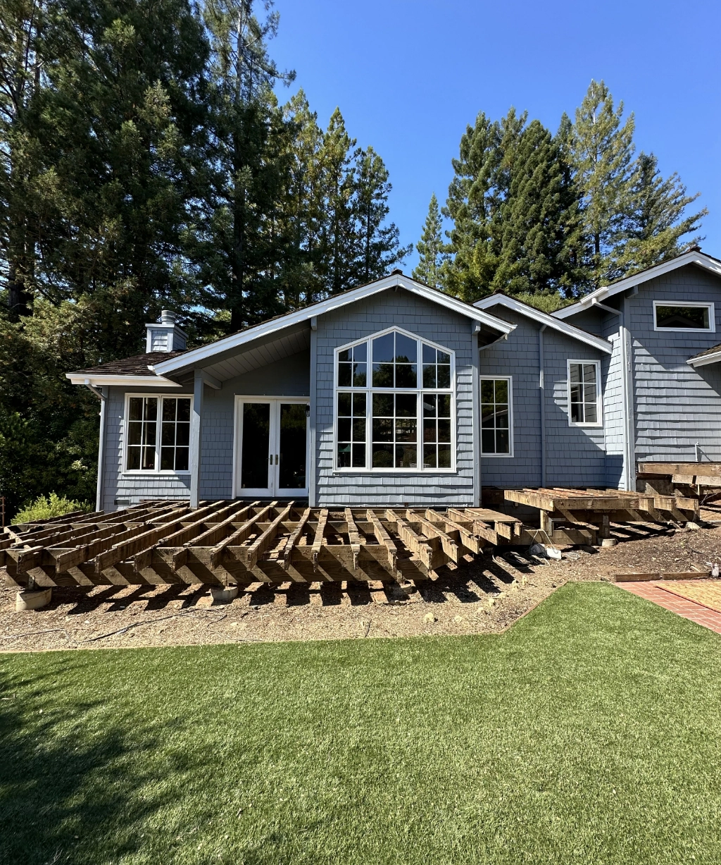 A blue house features large windows, surrounded by tall trees and an unfinished wooden deck frame. In the foreground, a green lawn stretches under a clear blue sky, creating a serene and picturesque setting.