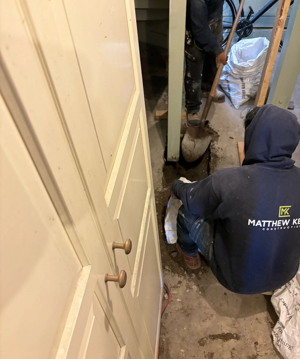 Two people in hoodies focus intently on their indoor construction task. One kneels with a tool in hand while the other stands nearby.