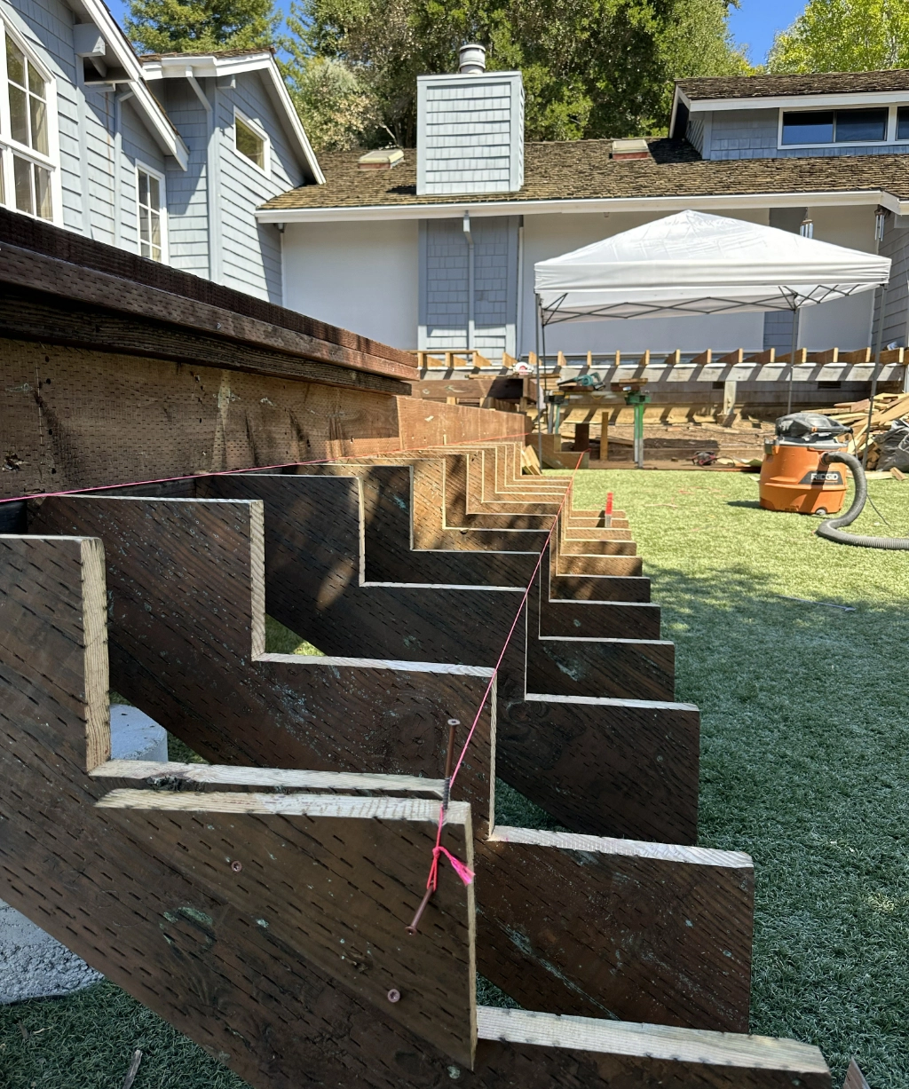 Wooden stair stringers are laid out in a backyard, aligned with a pink guide string. A partially constructed deck and house stand proudly in the background.