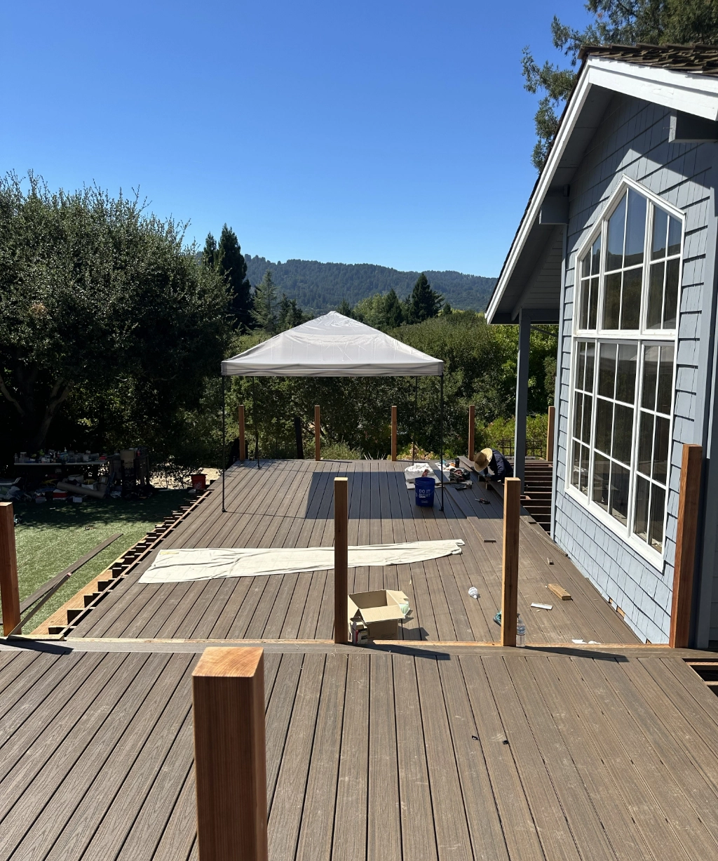 A wooden deck construction is underway beside a house. A canopy and scattered construction materials are visible, with trees and hills stretching in the background under a clear blue sky.