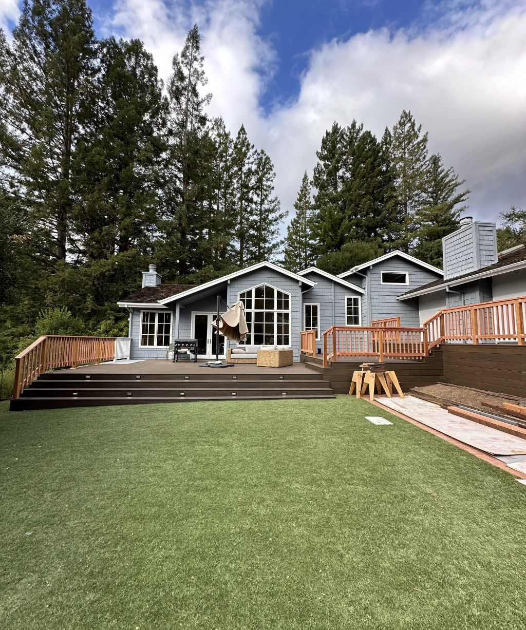 A modern gray house with large windows stands before tall trees. The backyard boasts a deck with wooden railings and patio furniture, all set on a grassy area under a partly cloudy sky.