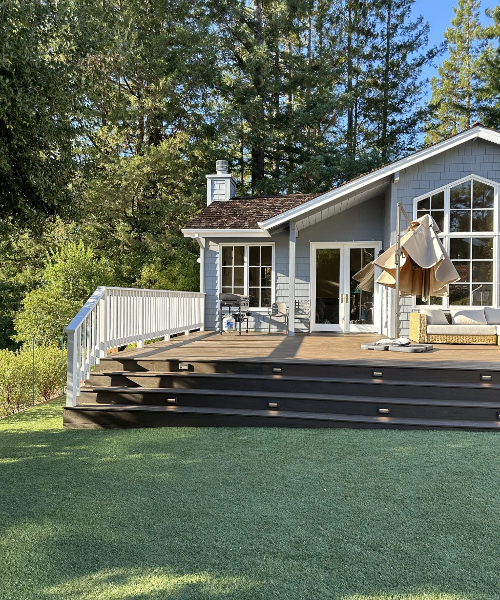 This modern house features a wooden deck, patio furniture, and large windows. It's beautifully nestled among trees and lush green grass, creating a serene oasis on a sunny day.