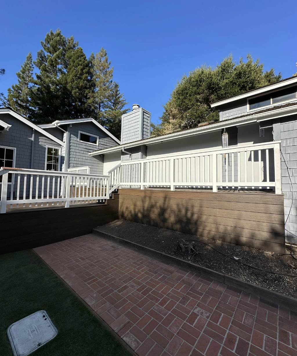 The rear view of this gray house features a white-railed wooden deck and a brick patio, all surrounded by trees under a clear blue sky.