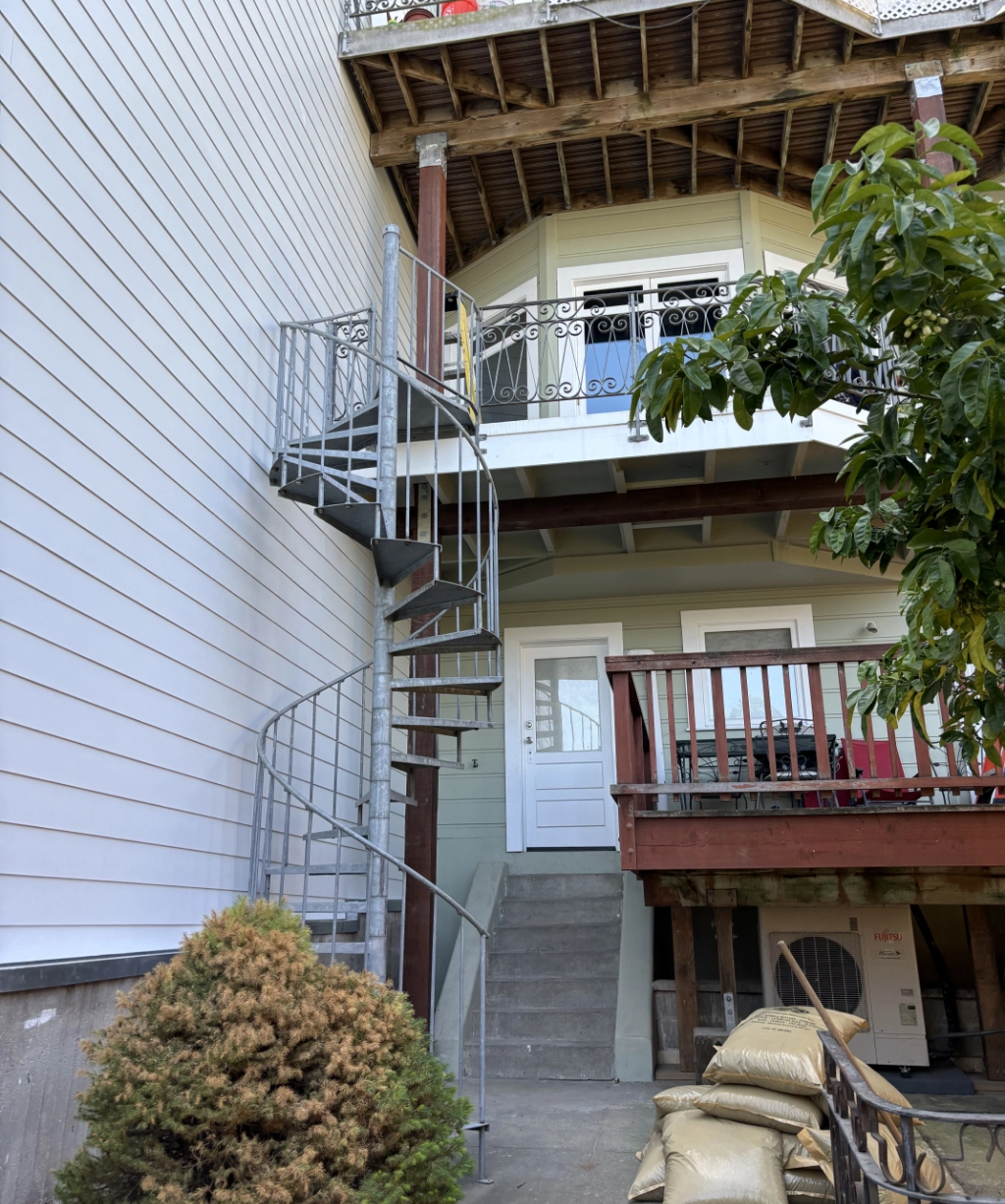 A metal spiral staircase elegantly winds up to an upper balcony of a multi-story house. There are stairs leading to a door, with several sandbags nestled near the base, adding a unique charm to this architectural gem.