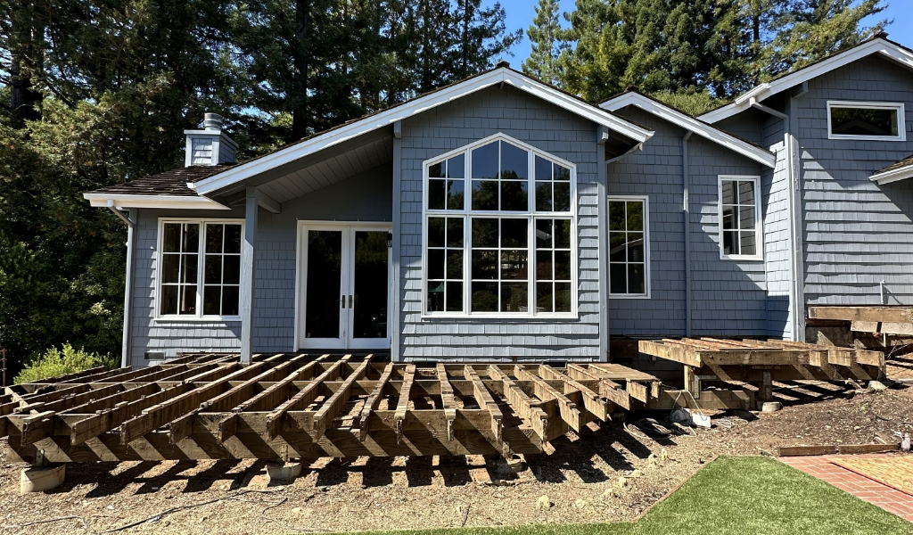 A house with gray siding and multiple large windows is surrounded by a wooden deck under construction in a tranquil wooded area.
