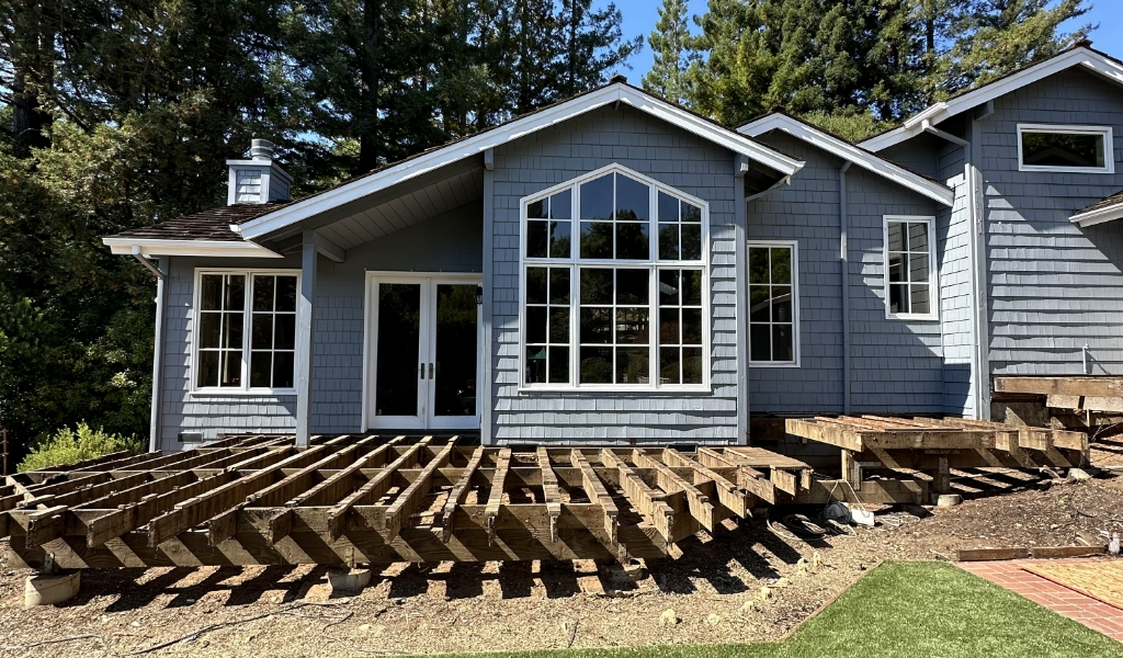 A house with gray siding and white trim stands serenely, showcasing an unfinished deck frame in the foreground, surrounded by a canopy of trees.