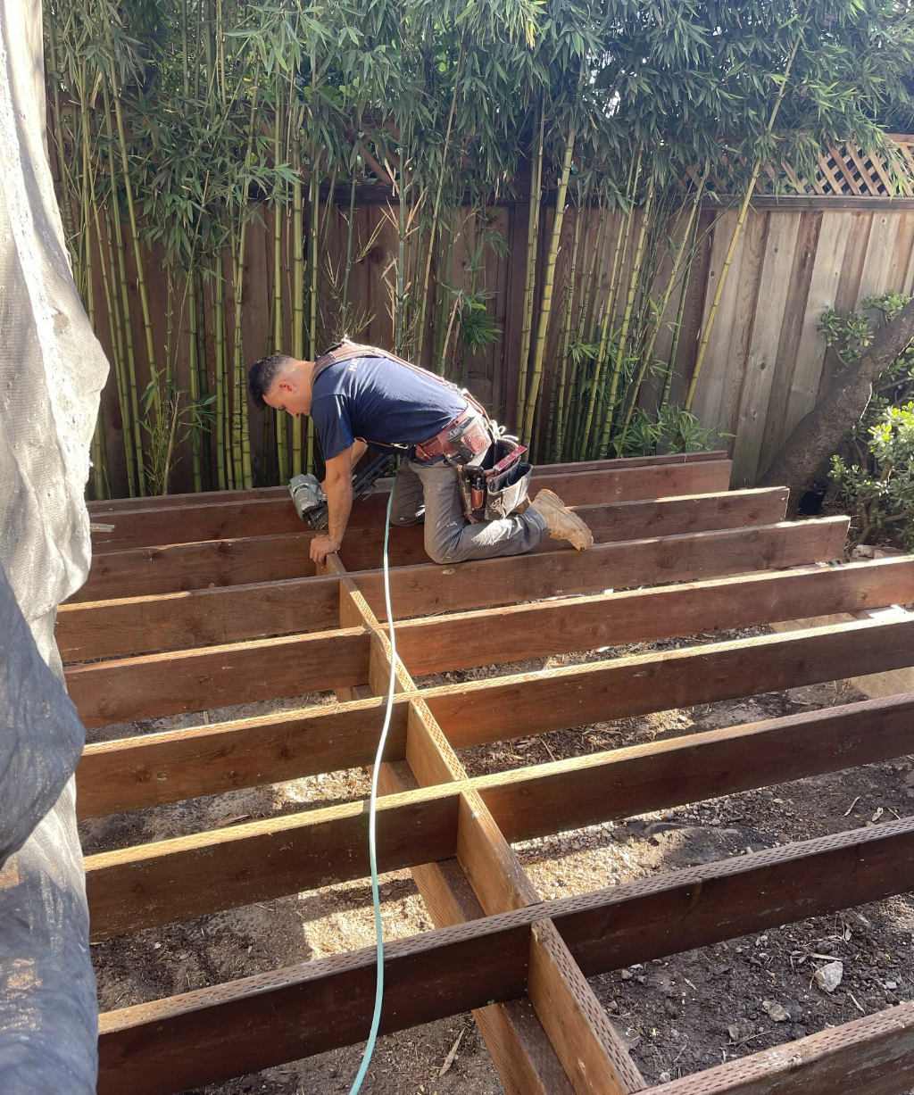 A person is kneeling and working on a wooden deck frame, surrounded by lush bamboo and a sturdy wooden fence.