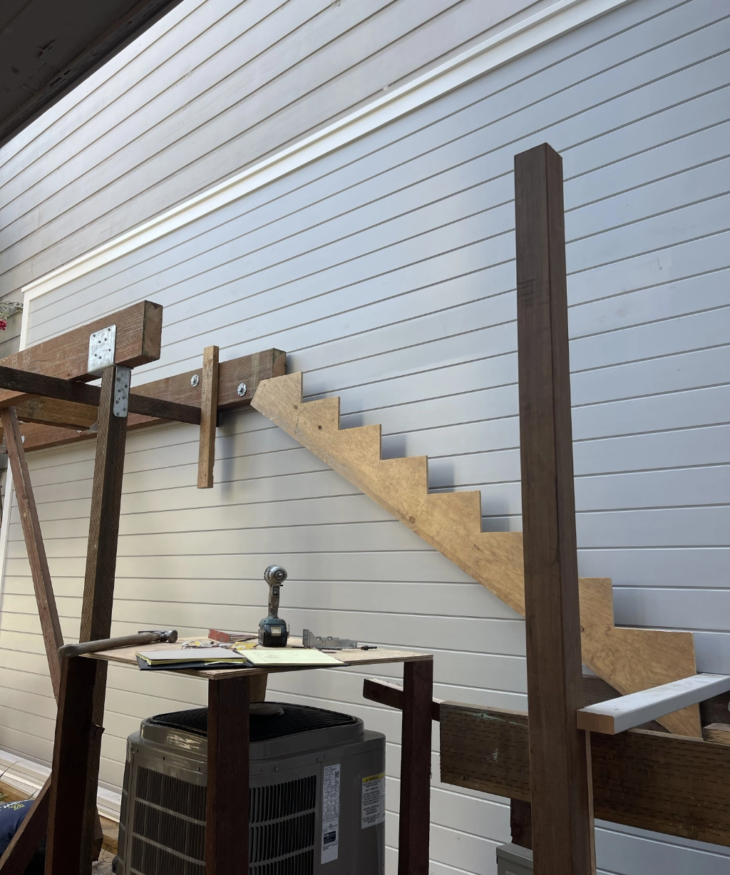 A wooden staircase in progress is attached to a wall, offering a glimpse into the development of a stylish residence. Nearby, tools and an air conditioning unit hint at the intricate work involved.