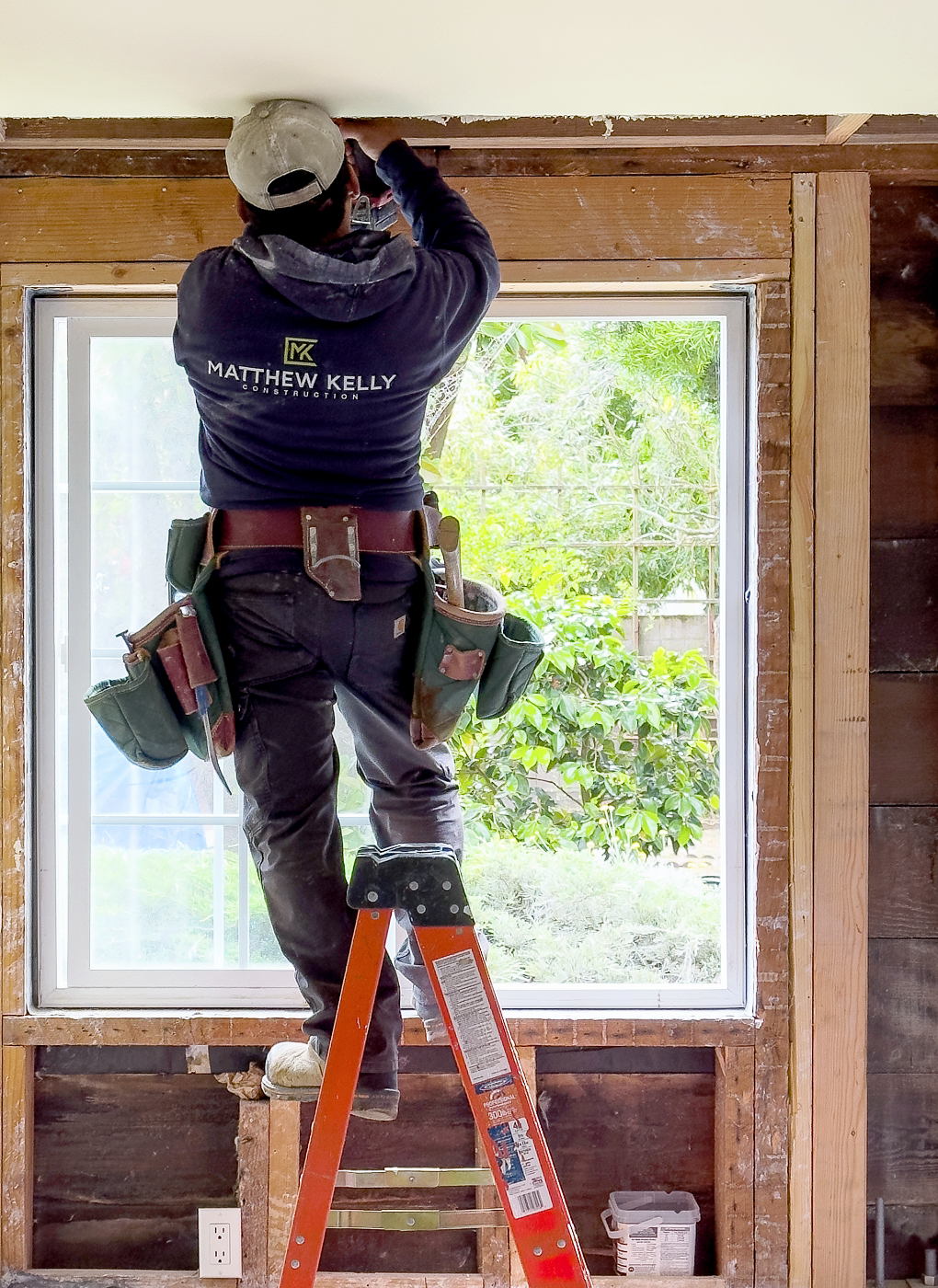 A worker from Matthew Kelly Construction stands on a ladder, expertly installing or repairing a window frame indoors. Outfitted with a tool belt and cap, he demonstrates the precision and quality synonymous with the Matthew Kelly brand.
