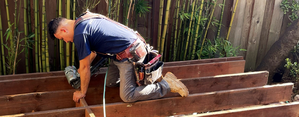 A Matthew Kelly Construction worker, donning a tool belt, kneels on wooden beams, expertly using a nail gun for construction work in a fenced outdoor area framed by bamboo plants.