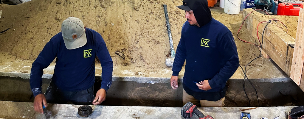 Two construction workers in blue shirts, part of the renowned Matthew Kelly Construction team, diligently labor at a concrete site. One bends over while the other stands holding tools, surrounded by sand and various construction materials.
